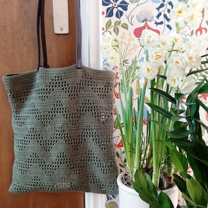 Op de afbeelding: Een groene gehaakte tote bag met bruine leren hengsels. De tas hangt aan een deur en er staan witte bloemen in een pot op de achtergrond.