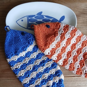 Peut inclure: Deux maniques en crochet en forme de poisson, l'une bleue et crème et l'autre orange et crème, avec un motif en point coquille texturé et une petite boucle sur le dessus pour suspendre, placées près d'une assiette ovale blanche avec un motif de poisson bleu.