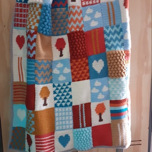Op de afbeelding: Een kleurrijk gebreid babydekentje met een patchwork ontwerp met harten, bomen, wolken en geometrische patronen. Het dekentje is gemaakt met verschillende texturen, waaronder bobbels en strepen.