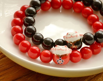 May include: Two bracelets, one with red beads and one with black beads, with a lucky cat charm on each. The bracelets are on a white plate.