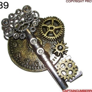 Può includere: Un ciondolo a forma di chiave in stile steampunk con una chiave d'argento, un quadrante in bronzo e vari ingranaggi in bronzo e argento. Il quadrante mostra i numeri 6, 7, 8 e 9. Il testo "LC39" è in alto a sinistra.