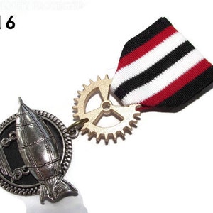 Könnte beinhalten: Eine silberne und goldene Steampunk-Anstecknadel mit einem Zahnrad und einem Luftschiff-Design. Die Nadel hat ein rot-weiß-schwarzes gestreiftes Band.