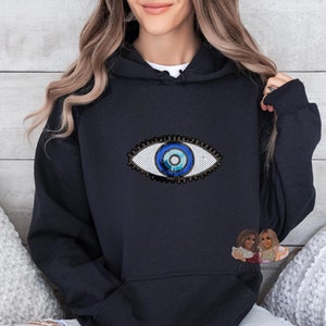 May include: A black hooded sweatshirt featuring a large, embroidered eye design. The eye is detailed with blue and white sequins, and a dark blue iris. The sweatshirt has a classic hoodie design with a front pocket and drawstring hood.