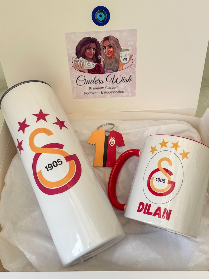 May include: A white gift box with a white tumbler, a white mug, and a red and yellow jersey keychain. The tumbler and mug feature a red and yellow logo with the text "1905" and five stars. The mug also has the name "DILAN" printed on it.