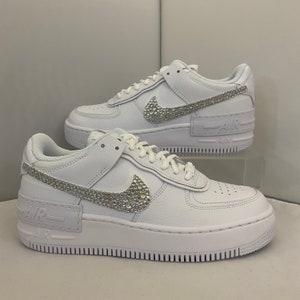 Puede incluir: Zapatillas blancas de cuero con suela de plataforma y un swoosh Nike incrustado de cristales.