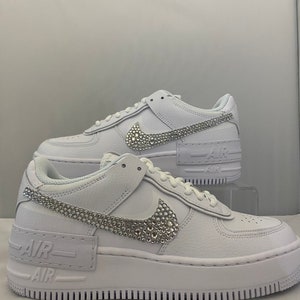 Puede incluir: Zapatillas bajas de cuero blanco con suela de plataforma blanca. El swoosh de Nike y el texto "Air" en el lateral de la zapatilla est&aacute;n cubiertos de pedrer&iacute;a.