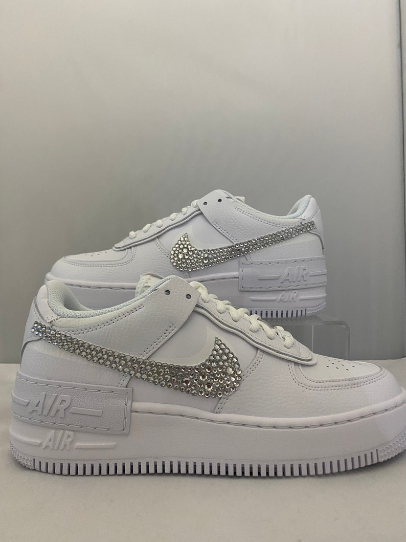 Puede incluir: Zapatillas bajas de cuero blanco con suela de plataforma blanca. El swoosh de Nike y el texto "Air" en el lateral de la zapatilla est&aacute;n cubiertos de pedrer&iacute;a.