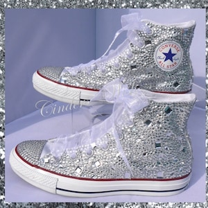 May include: White high-top Converse sneakers covered in silver glitter and rhinestones. The Converse logo is visible on the side of the shoe.