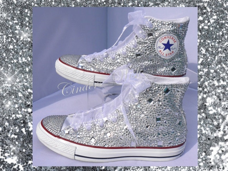 May include: White high-top Converse sneakers covered in silver glitter and rhinestones. The Converse logo is visible on the side of the shoe.