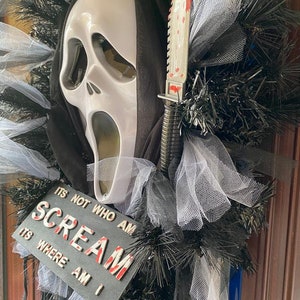 May include: A Halloween wreath with a white ghost mask, a black hooded robe, and a bloody knife. The wreath is decorated with black and white tulle and black tinsel. The wreath has a sign that reads "It's not who am I, it's where am I, Scream."
