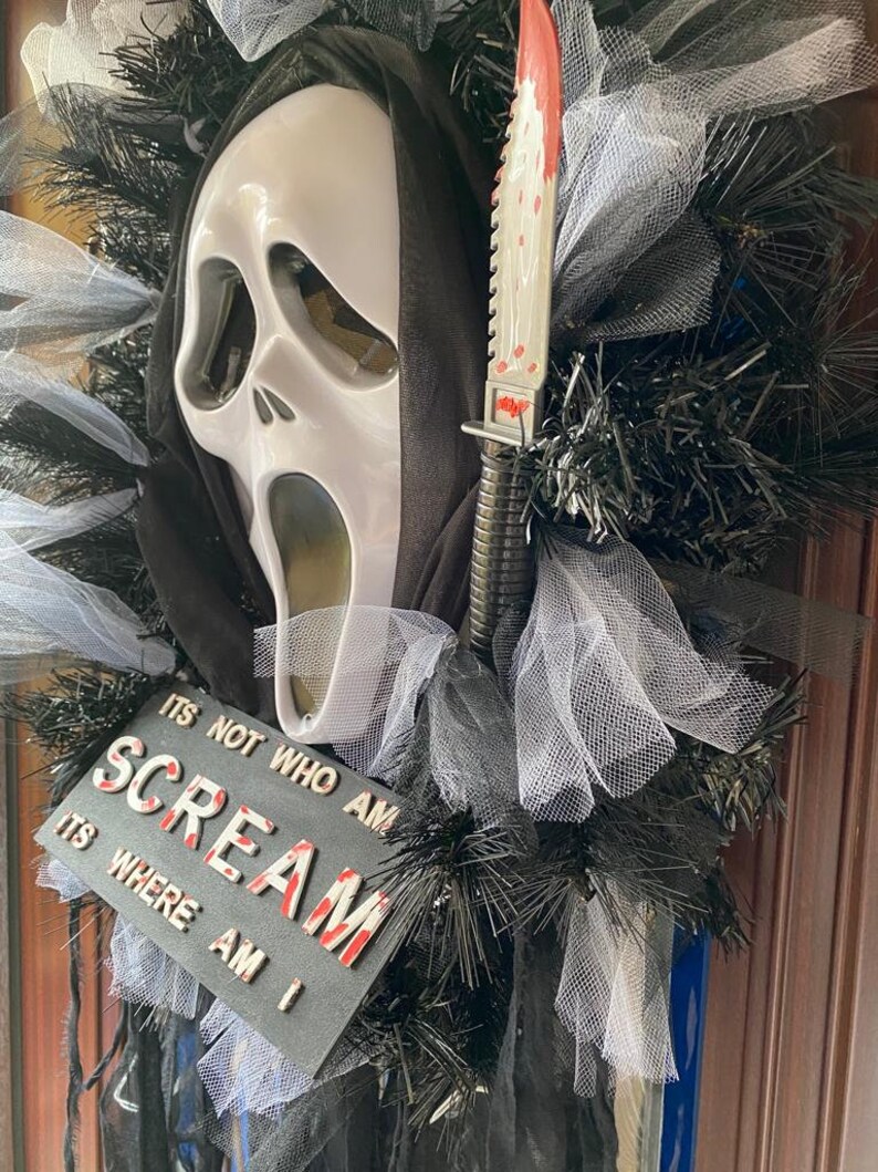 May include: A Halloween wreath with a white ghost mask, a black hooded robe, and a bloody knife. The wreath is decorated with black and white tulle and black tinsel. The wreath has a sign that reads "It's not who am I, it's where am I, Scream."
