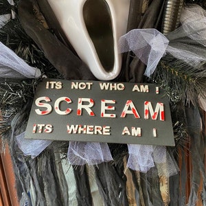 May include: A black and white sign with the words "It's not who am I, Scream, It's where am I" in white letters with red splatters. The sign is decorated with black and white tulle and a white ghost mask with a black mouth.