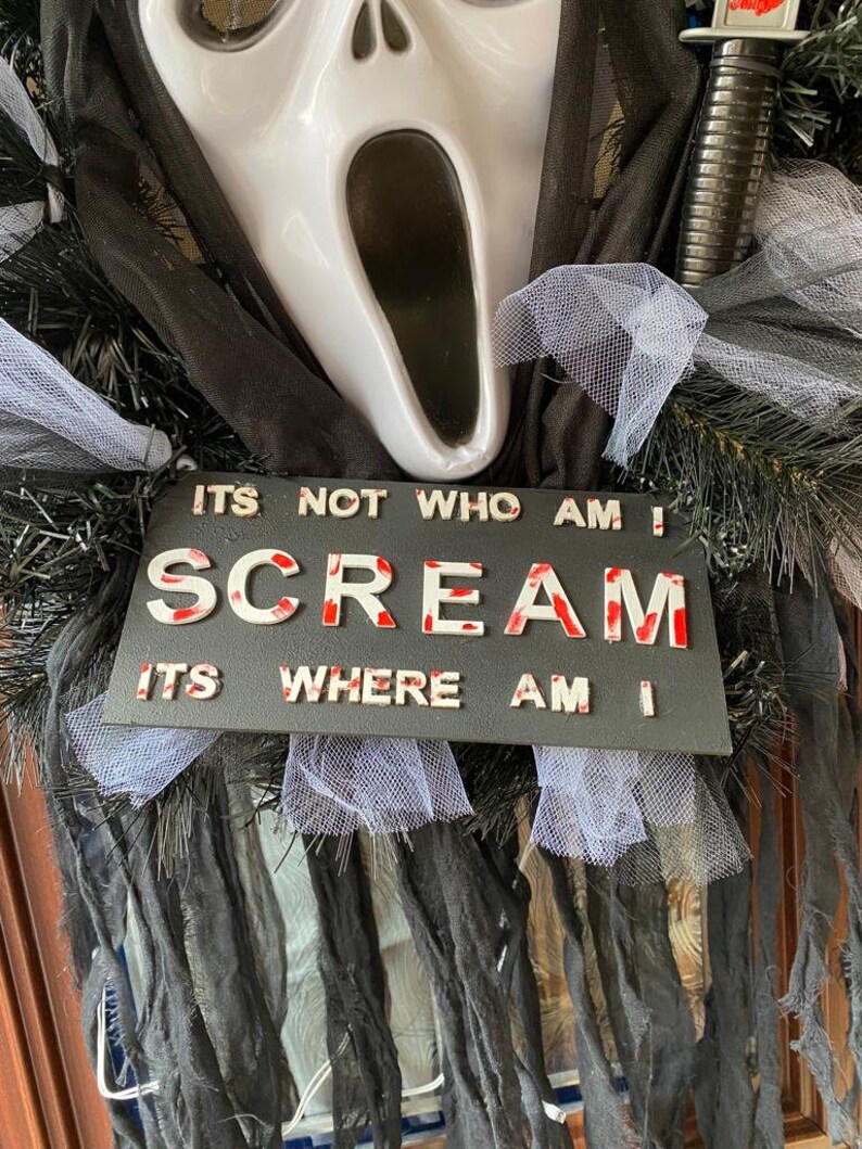 May include: A black and white sign with the words "It's not who am I, Scream, It's where am I" in white letters with red splatters. The sign is decorated with black and white tulle and a white ghost mask with a black mouth.