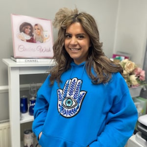 May include: A bright blue hoodie featuring a large embroidered patch of a hamsa hand with a blue eye in the centre. The hamsa hand is surrounded by blue and white sequins.