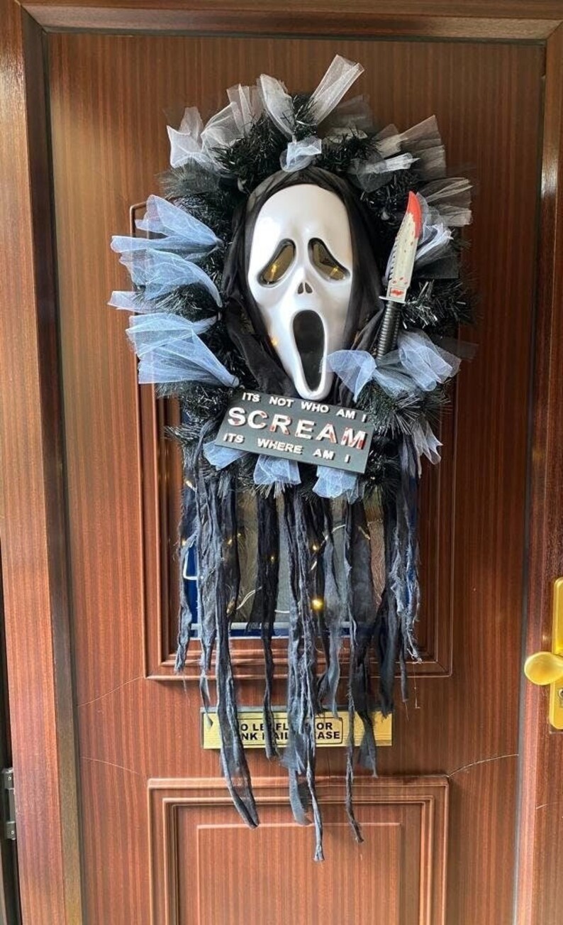 May include: A Halloween door wreath with a white ghost mask, black and blue tulle, and a sign that reads "It's not who am I Scream It's where am I".