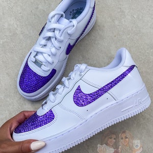May include: White leather sneakers with a purple rhinestone swoosh and toe. The sneakers have a white sole and a white lace.