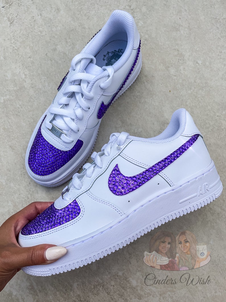 May include: White leather sneakers with a purple rhinestone swoosh and toe. The sneakers have a white sole and a white lace.