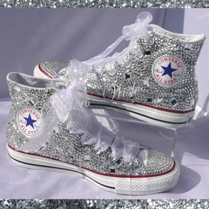 May include: White high-top Converse sneakers covered in silver glitter and rhinestones. The Converse logo and All Star text are visible on the side of the shoes.