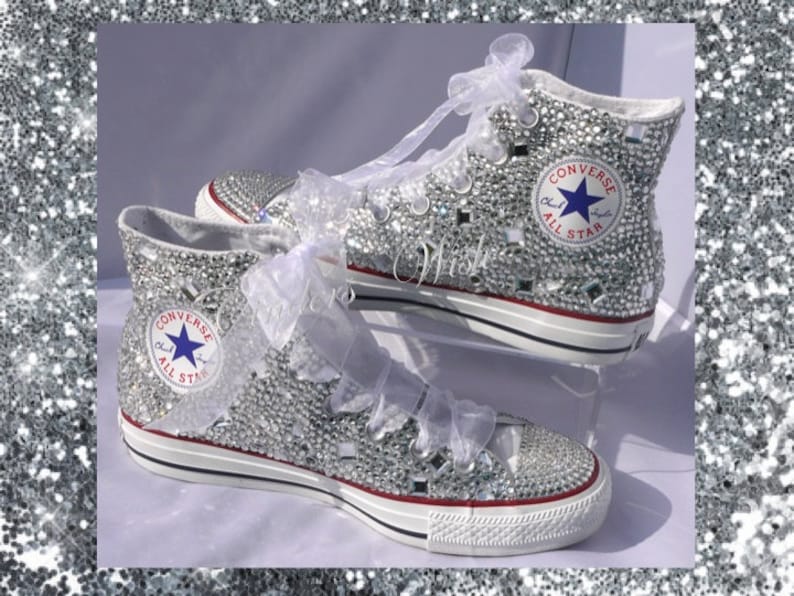 May include: White high-top Converse sneakers covered in silver glitter and rhinestones. The Converse logo and All Star text are visible on the side of the shoes.