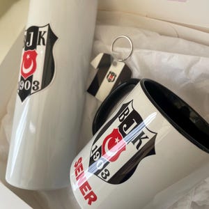 May include: A white mug with a black handle and a black and white logo with a red crescent moon and star and the text "BJK 1903" and the word "SELER" in red. The mug is next to a white tumbler with the same logo. A keychain with the same logo is also included.