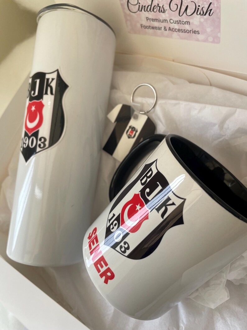 May include: A white mug with a black handle and a black and white logo with a red crescent moon and star and the text "BJK 1903" and the word "SELER" in red. The mug is next to a white tumbler with the same logo. A keychain with the same logo is also included.