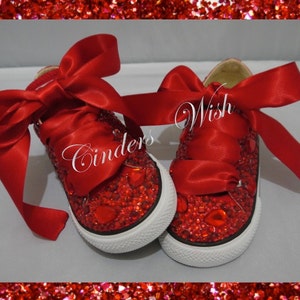Puede incluir: Zapatillas Converse rojas brillantes con lazos de cinta de satén rojo. Las zapatillas están decoradas con diamantes de imitación rojos y el texto "Wish" es visible en una zapatilla.