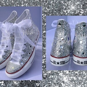 May include: White high-top sneakers covered in silver glitter and rhinestones. The sneakers have white laces and a white ribbon tied around the ankles. The words "All Star" are printed on the back of each shoe.