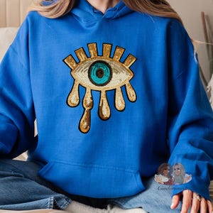May include: A royal blue hoodie featuring a gold sequin eye design. The eye has a turquoise blue pupil and gold eyelashes. The eye is dripping with gold sequins.