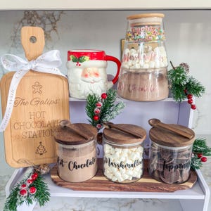 May include: A festive hot chocolate station featuring a wooden board, Santa mug, and glass jars with toppings. The board reads "The Galips Hot Chocolate Station". Jars contain hot chocolate mix, marshmallows, and chocolate toppings, all arranged on a white shelf.