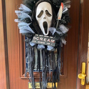 May include: A Halloween door wreath with a white ghost mask, black tulle, and a red-handled knife. The wreath has a sign that reads "ITS NOT WHO AM I SCREAM ITS WHERE AM I".