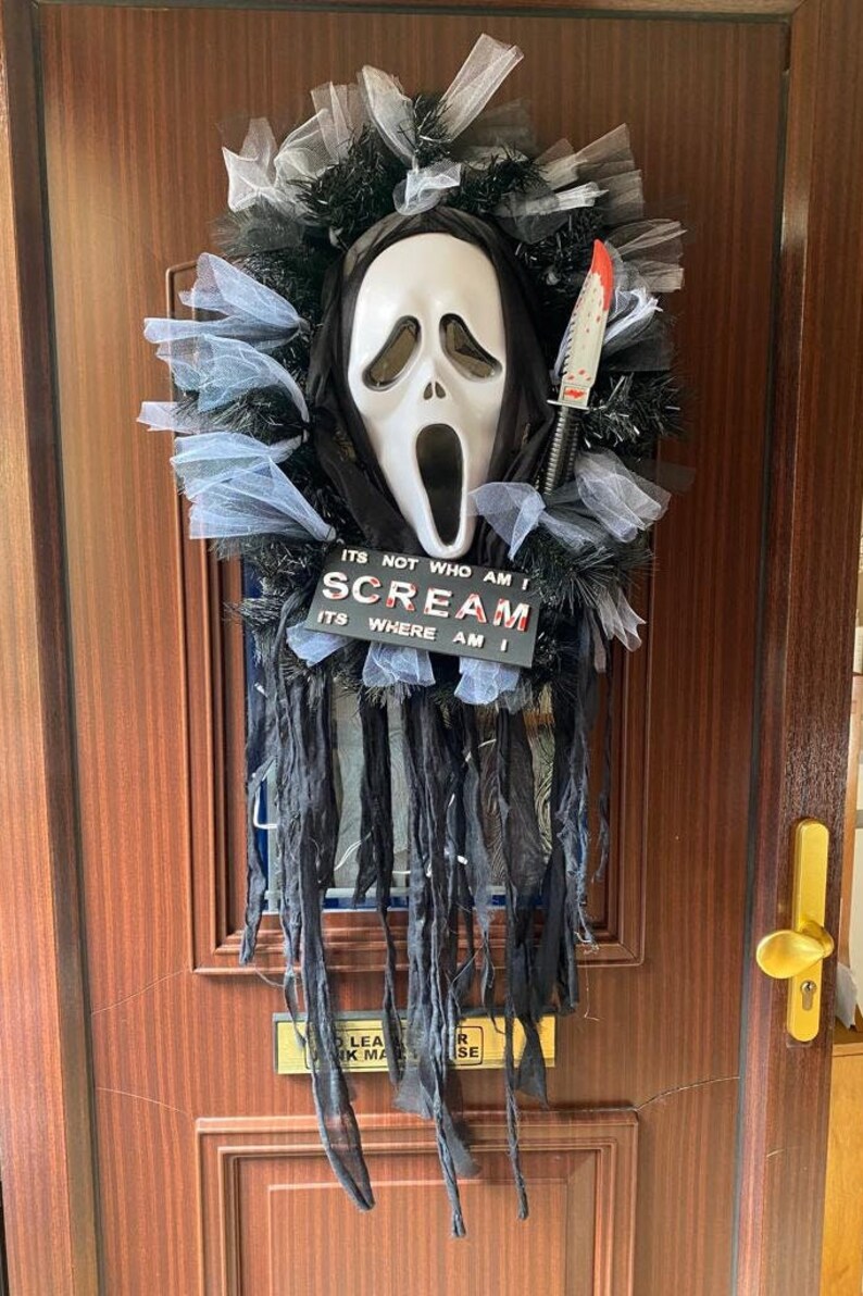 May include: A Halloween door wreath with a white ghost mask, black tulle, and a red-handled knife. The wreath has a sign that reads "ITS NOT WHO AM I SCREAM ITS WHERE AM I".