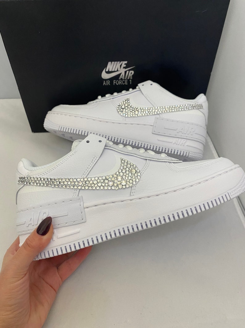 Puede incluir: Zapatillas Nike Air Force 1 de cuero blanco con un swoosh adornado con cristales y una banda adornada con cristales en la parte delantera de la zapatilla. Las zapatillas est&aacute;n en una caja con el logotipo de Nike y las palabras "Air Force 1" en ella.