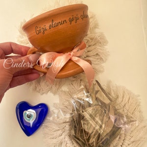 May include: A terracotta cup with a pink ribbon and the text "Gözü olanın gözü çıksın" in black. The cup is sitting on a white fluffy surface. There is a blue glass heart with a white and blue evil eye charm and a bag of dried herbs on the surface.