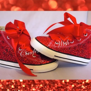 Puede incluir: Zapatillas rojas de caña alta cubiertas de purpurina roja con un lazo de satén rojo. Las zapatillas tienen una suela blanca y las palabras "Wish" están bordadas en el lateral.
