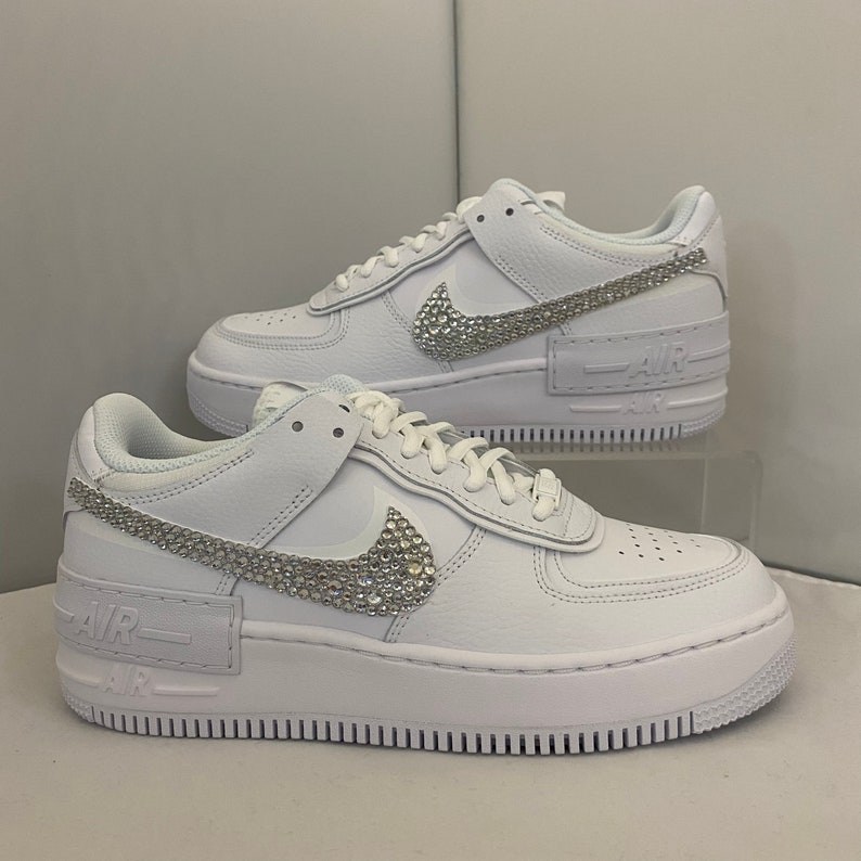 Puede incluir: Zapatillas bajas de cuero blanco con suela de plataforma. El swoosh de Nike est&aacute; adornado con pedrer&iacute;a. El texto "AIR" est&aacute; impreso en el lateral de la zapatilla.