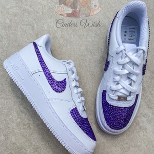 May include: White leather low-top sneakers with purple rhinestone accents on the swoosh logo and toe. The sneakers have a white rubber sole and a white lace-up closure.