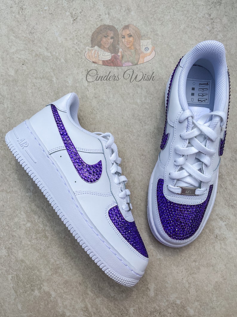May include: White leather low-top sneakers with purple rhinestone accents on the swoosh logo and toe. The sneakers have a white rubber sole and a white lace-up closure.