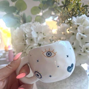May include: A white ceramic mug with a dimpled, uneven surface texture. The mug features a repeating pattern of dark blue and gold evil eye symbols, small dark blue stars, and a dark blue heart icon with a white eye graphic inside.