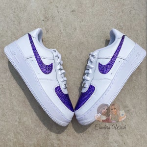May include: White leather low-top sneakers with a purple rhinestone swoosh and a purple rhinestone toe. The sneakers are on a white background.