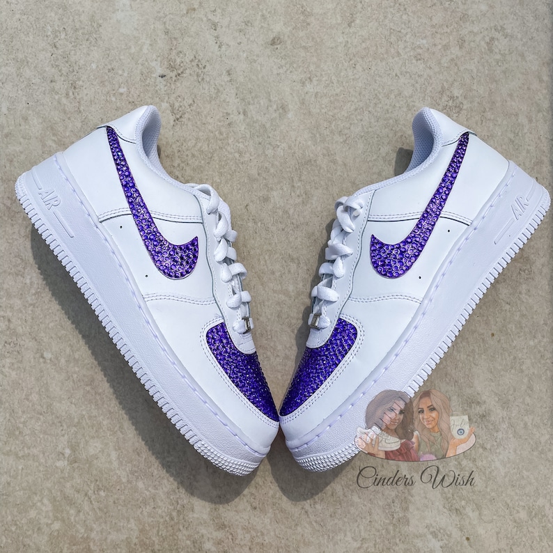 May include: White leather low-top sneakers with a purple rhinestone swoosh and a purple rhinestone toe. The sneakers are on a white background.