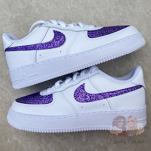 May include: White low-top sneakers with a purple rhinestone Nike swoosh. The sneakers have a white sole and a white leather upper.