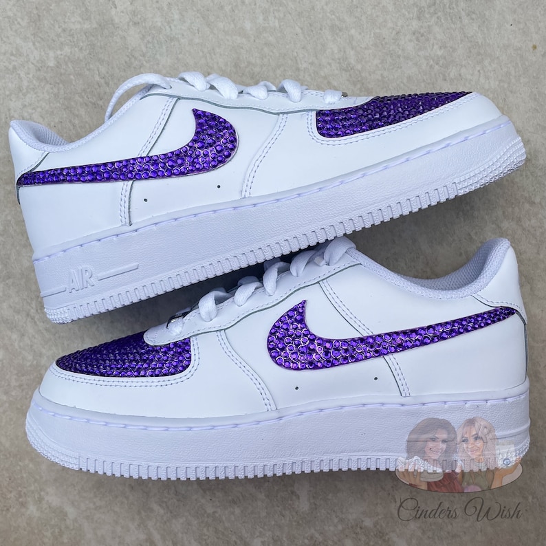 May include: White low-top sneakers with a purple rhinestone Nike swoosh. The sneakers have a white sole and a white leather upper.