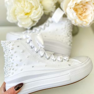 Puede incluir: Zapatillas altas blancas adornadas con perlas. Las zapatillas tienen cordones blancos y una suela blanca gruesa. La parte superior del zapato está cubierta de pequeñas perlas. Cintas de satén blancas atadas con un lazo.