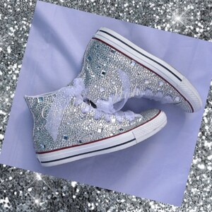 May include: A pair of white high-top sneakers covered in silver glitter and rhinestones. The sneakers have white laces and a white ribbon tied around the ankle.