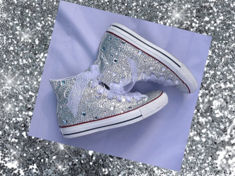 May include: A pair of white high-top sneakers covered in silver glitter and rhinestones. The sneakers have white laces and a white ribbon tied around the ankle.