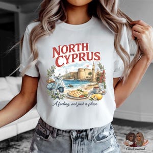 May include: A white t-shirt featuring a graphic print with the text NORTH CYPRUS and A feeling, not just a place. The illustration depicts a blue scooter, a coastal fortress, a plate of grilled food, a cup of tea, and lemons surrounded by greenery.
