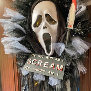 May include: A Halloween wreath featuring a white Ghostface mask with a black hood, a bloody knife, and a black sign that reads "ITS NOT WHO AM I SCREAM ITS WHERE AM I".