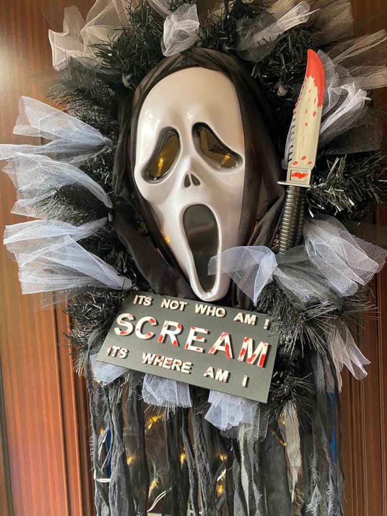 May include: A Halloween wreath featuring a white Ghostface mask with a black hood, a bloody knife, and a black sign that reads "ITS NOT WHO AM I SCREAM ITS WHERE AM I".