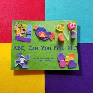 May include: A green book with the title "ABC, CAN YOU FIND ME?" written in white. The book has a variety of colorful felt shapes and letters glued to the cover, including a blue bird, a red heart, and a yellow star.