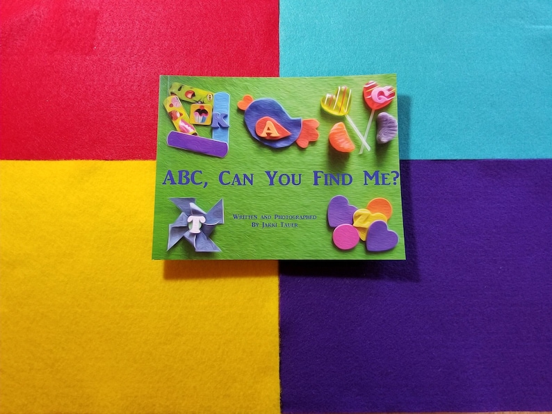 May include: A green book with the title "ABC, CAN YOU FIND ME?" written in white. The book has a variety of colorful felt shapes and letters glued to the cover, including a blue bird, a red heart, and a yellow star.